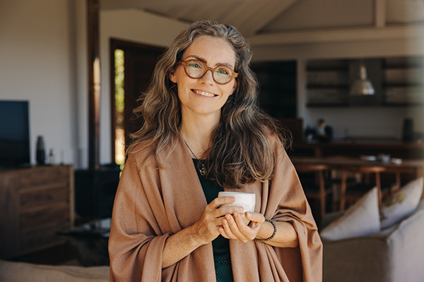 A 40-something white woman in casual dress and glasses holds a cup of coffee and smiles at the viewer.